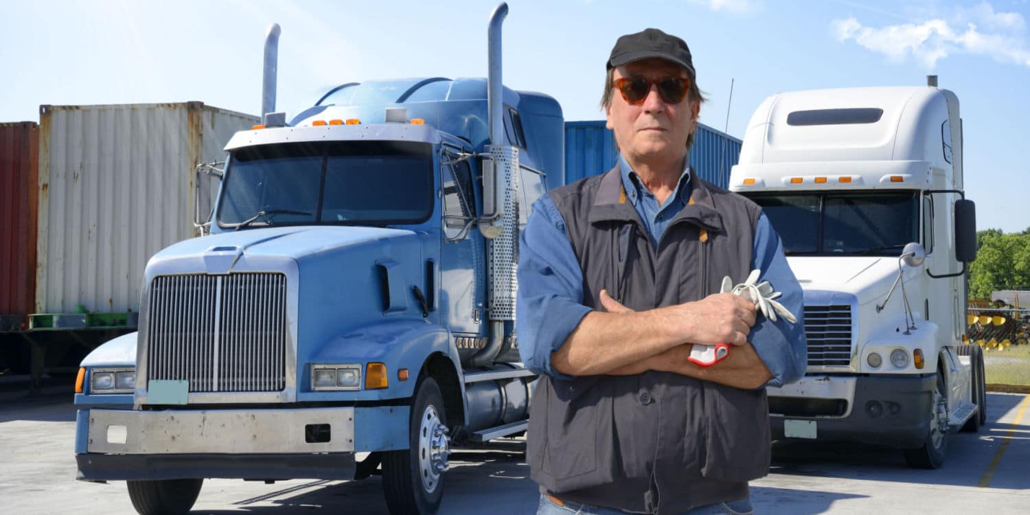 A man standing with his arms crossed in front of two semi-trucks representing an owner-operater vs company driver