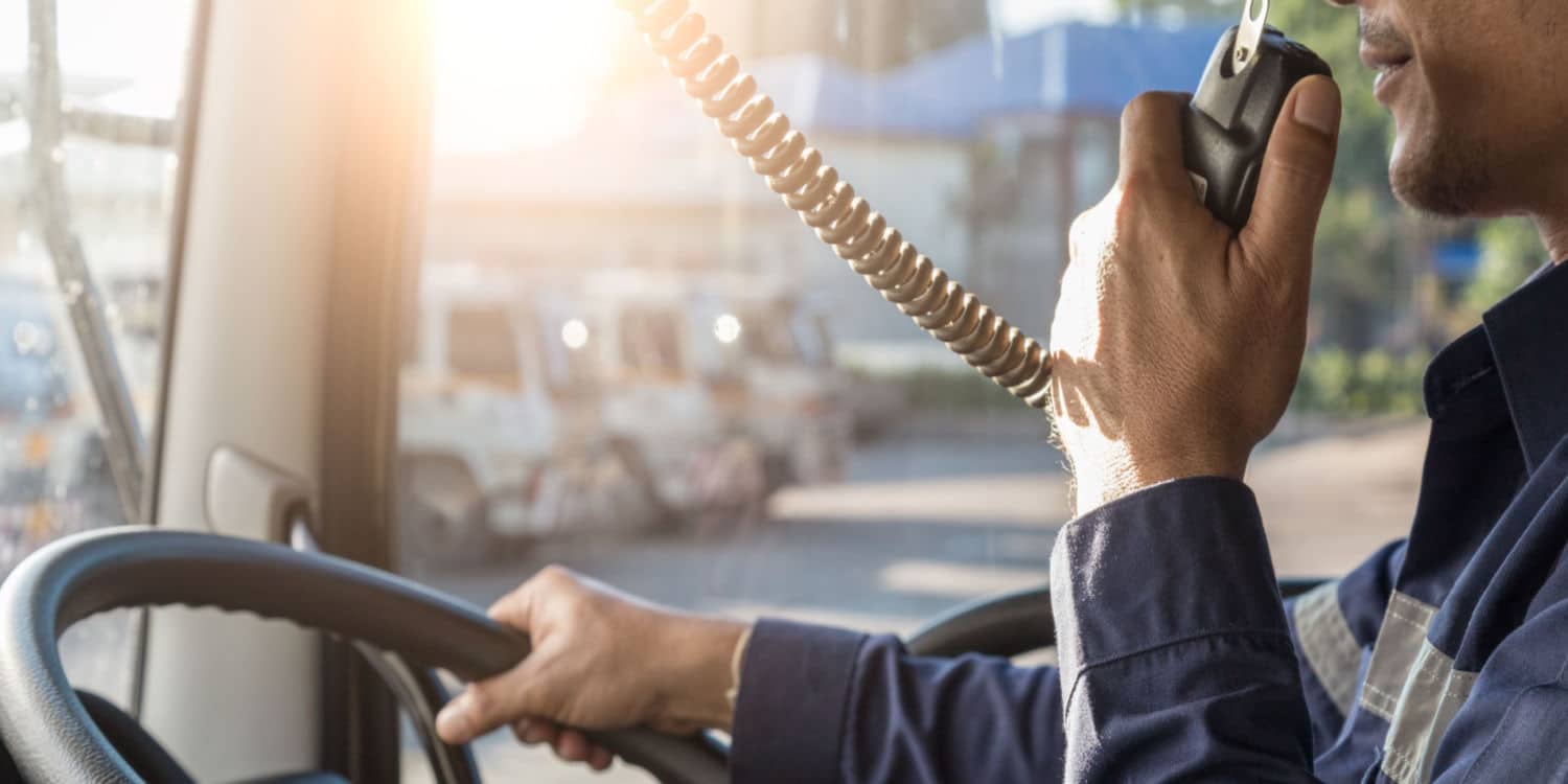 A close up of a trucker holding a radio in one hand and the steering wheel in the other as they speak to their driver dispatcher