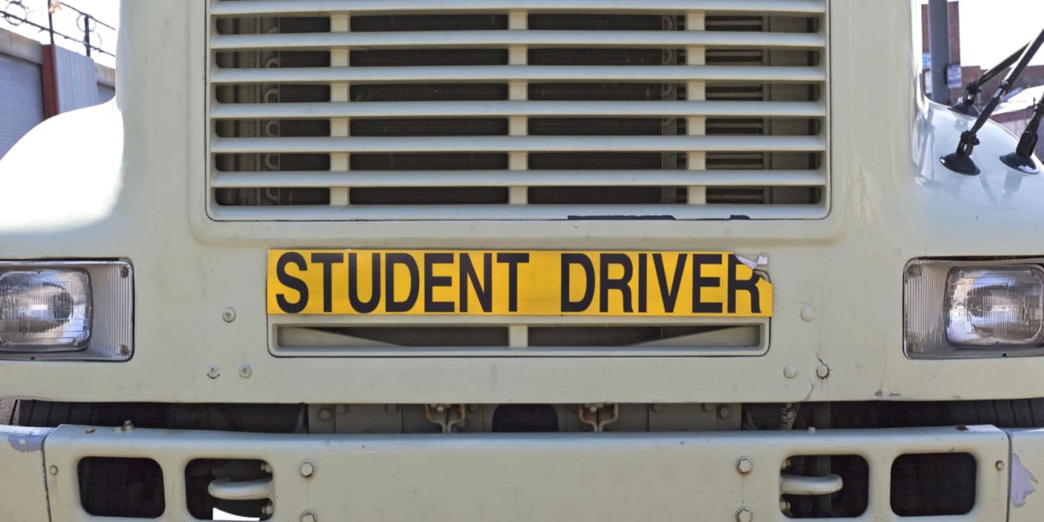 A yellow sign on the front of a semi-truck that reads student driver signifying CDL school challenges
