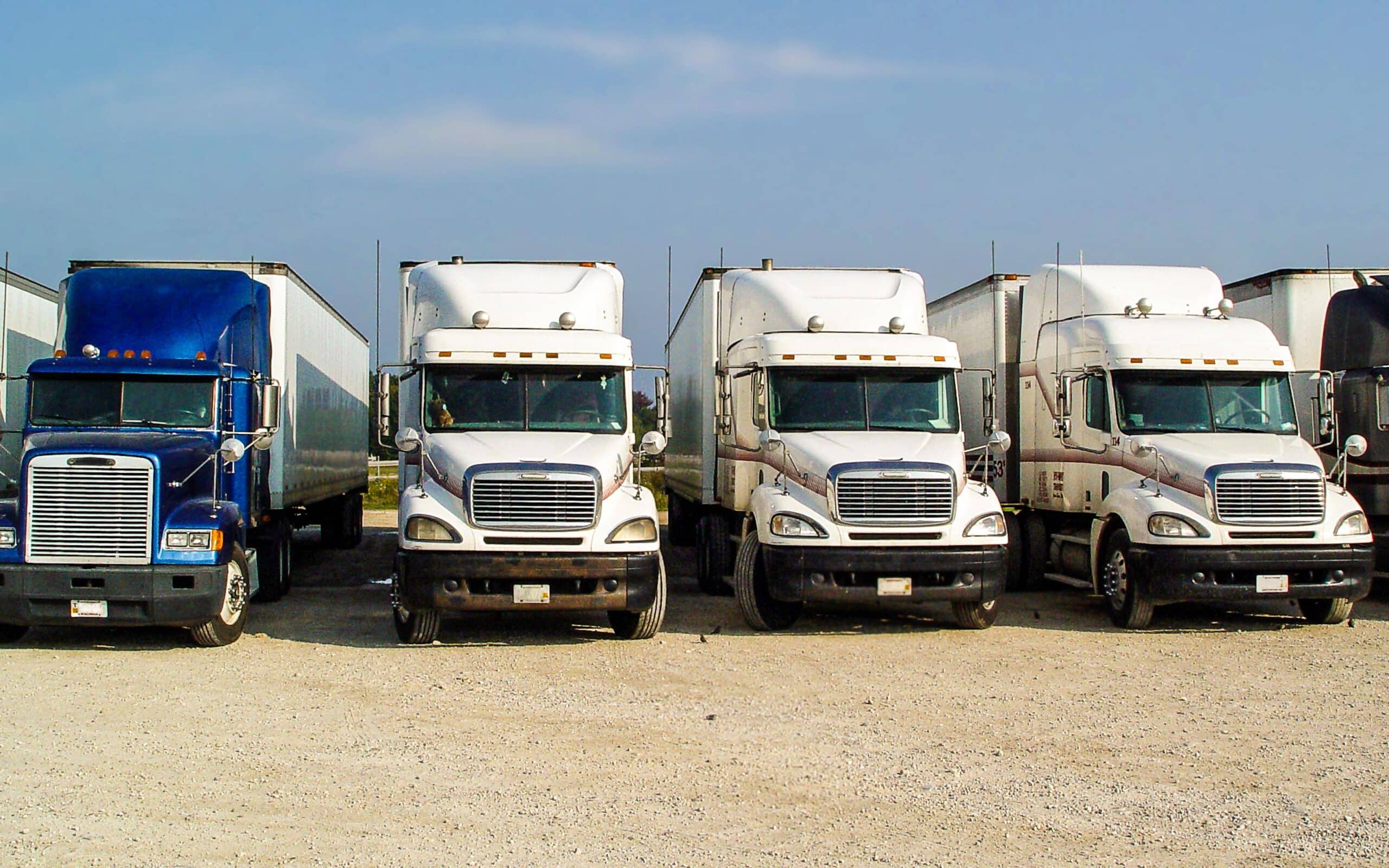 A row of five semi-trucks parked on a gravel lot, with the first truck being blue and the remaining four trucks being white.