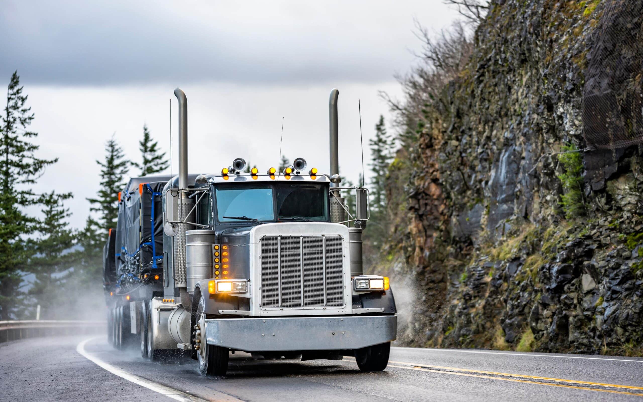 A large semi-truck with a silver front grille and multiple lights on top is driving on a wet, winding mountain road. The truck is pulling a blue trailer and there are trees and rocky cliffs in the background.