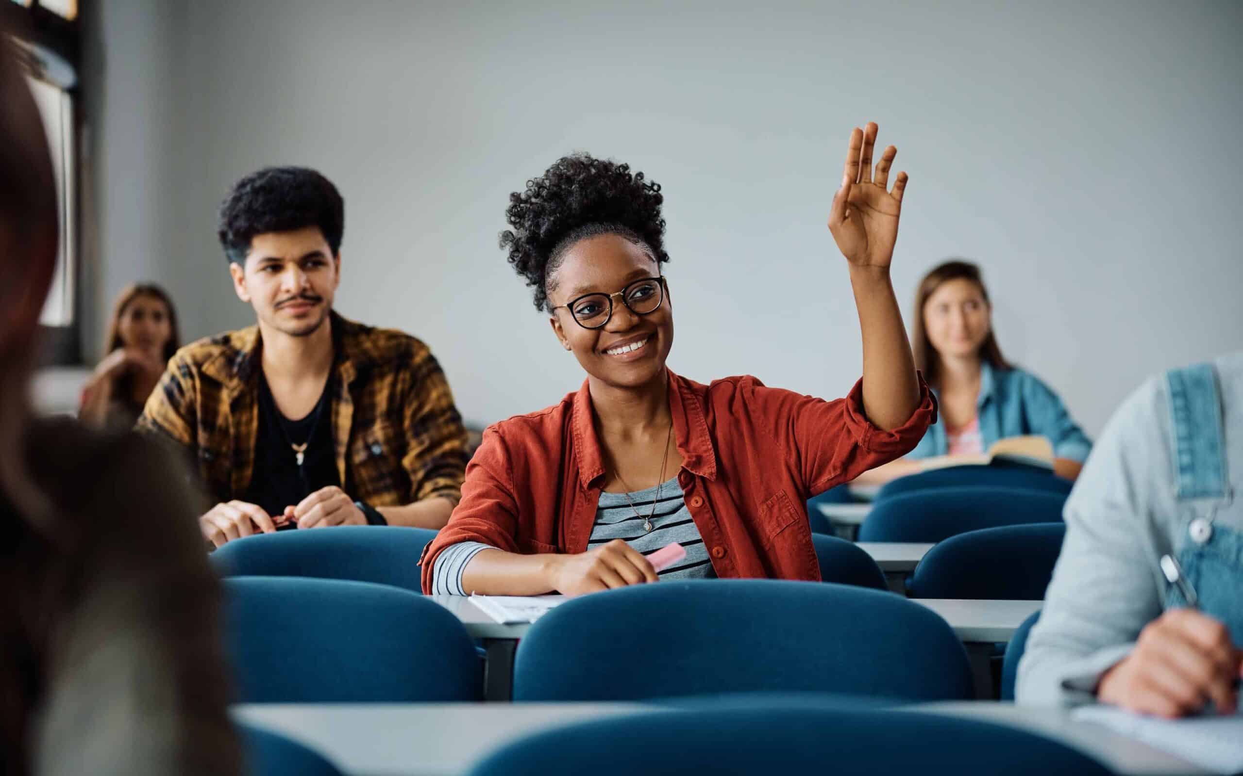 Student in a red shirt raising a hand in a classroom with blue chairs and desks, while others are seated and engaged in writing or reading