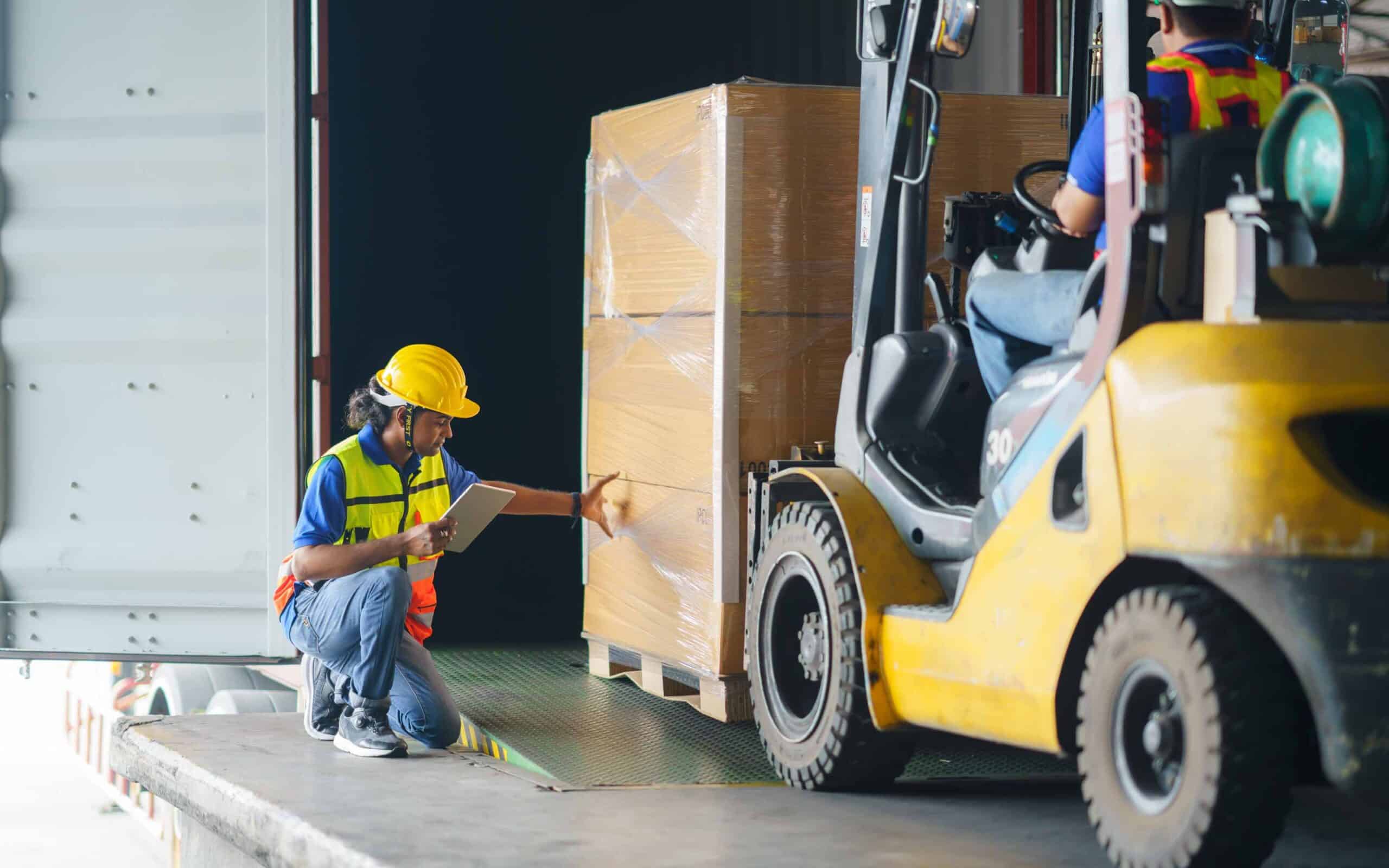 Two warehouse workers handling a large plastic-wrapped pallet—one operating a yellow forklift, the other kneeling with a tablet, both wearing safety vests and helmets in a loading dock setting