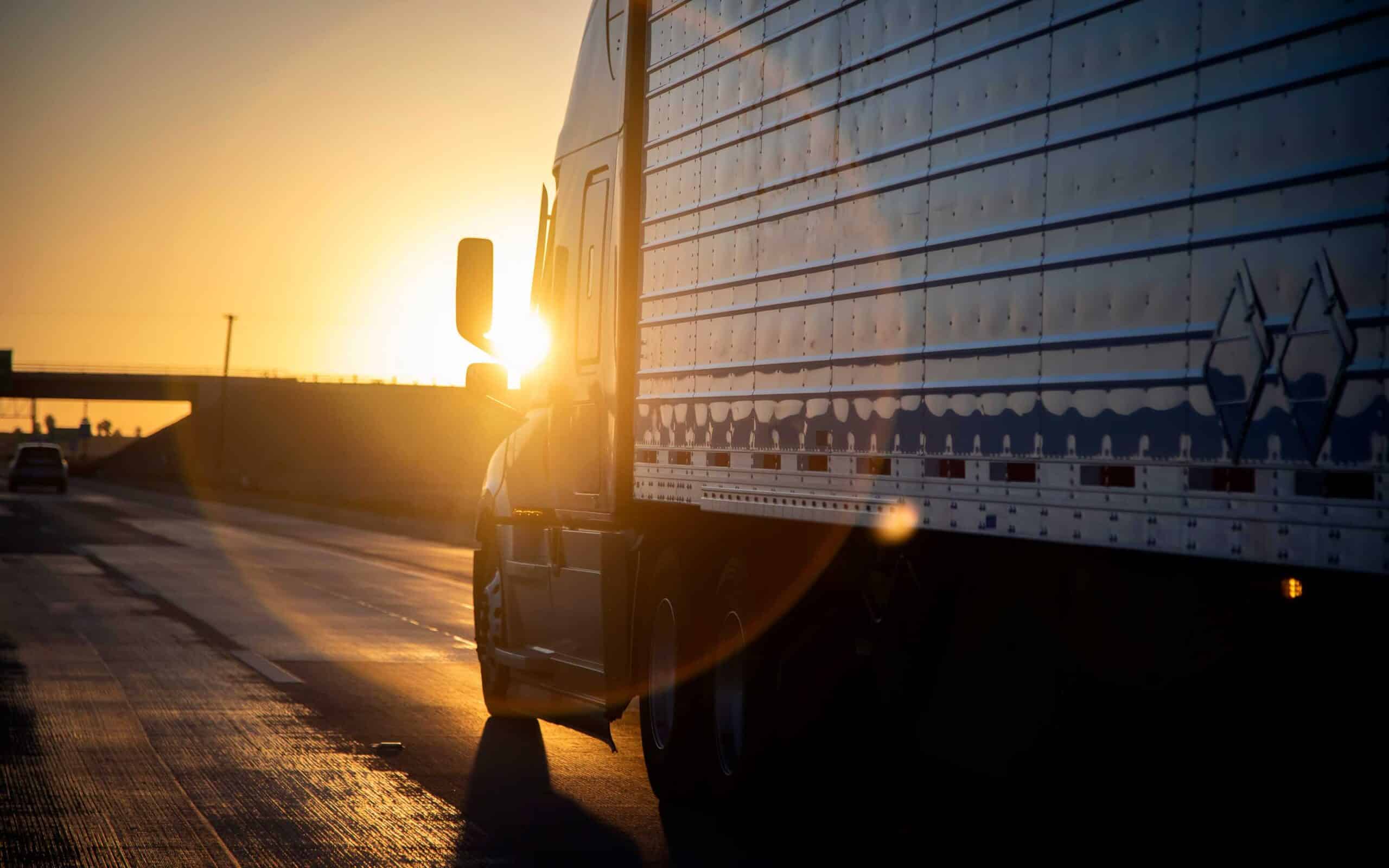 A large semi-truck drives along an empty highway during sunset, with golden light casting dramatic shadows and highlighting the truck’s reflective trailer.