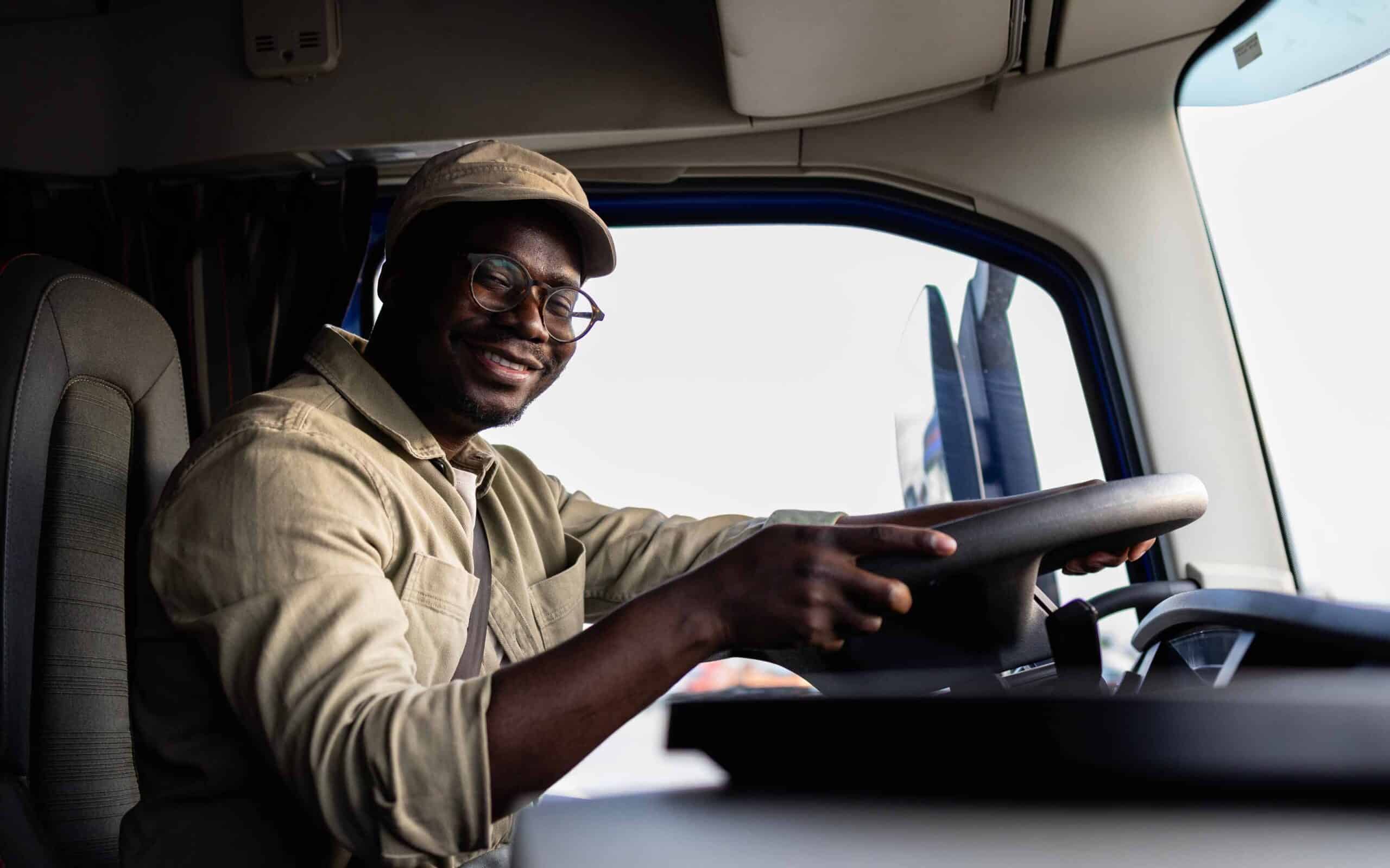 A happy truck driver wearing glasses and a cap smiles while holding the steering wheel, ready to start his truck driving career.